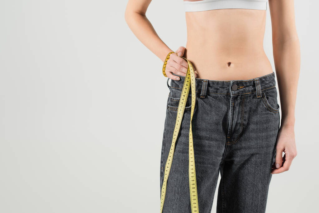 oříznutý pohled na fit ženu v džínách stojící s rukou na boku a měřicí pásky izolované na šedé - Fotografie, Obrázek