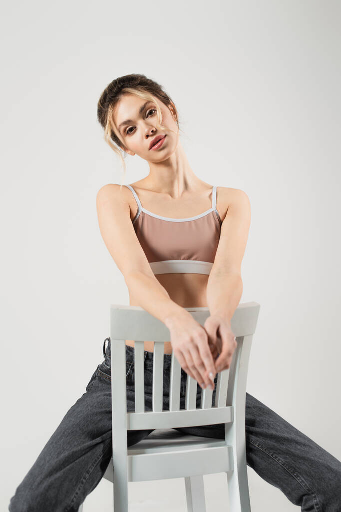 λεπτή γυναίκα με φυσικό μακιγιάζ κάθεται στην καρέκλα και κοιτάζοντας κάμερα απομονωμένη σε γκρι - Φωτογραφία, εικόνα