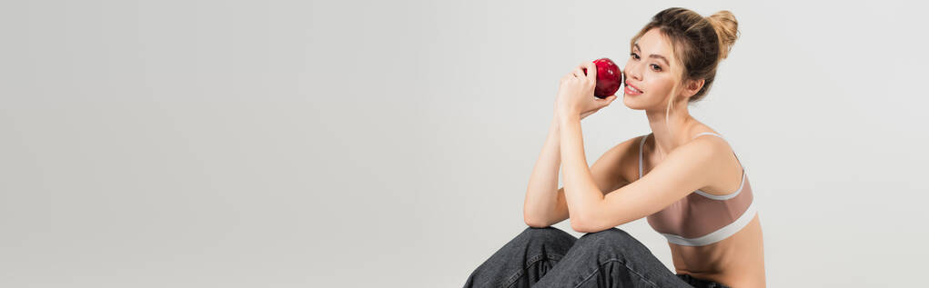 positiivinen nuori nainen urheilu top istuu kypsä omena eristetty harmaa, banneri - Valokuva, kuva