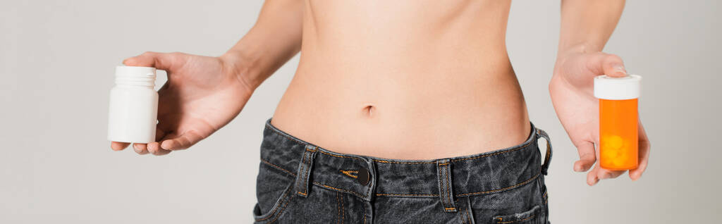 gedeeltelijk zicht op fit vrouw in jeans met vitamines in containers geïsoleerd op grijs, banner - Foto, afbeelding
