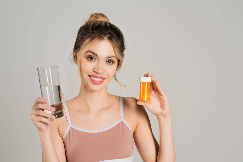 žena s dokonalou pletí s úsměvem na kameru při držení sklenice vody a vitamínů izolovaných na šedé - Fotografie, Obrázek