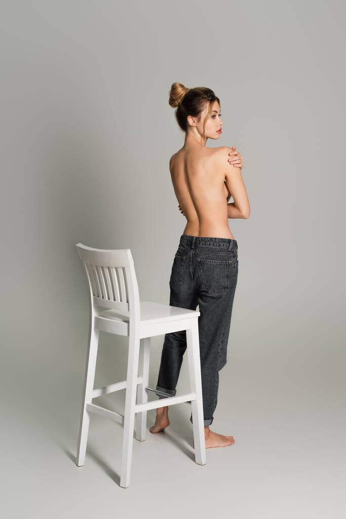 半裸の女性のジーンズ姿が白い椅子の近くに立って灰色の背景を見ています - 写真・画像