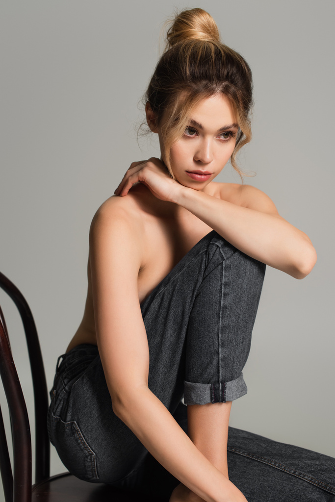 young half naked woman in jeans sitting on wooden chair and looking away isolated on grey - Photo, Image