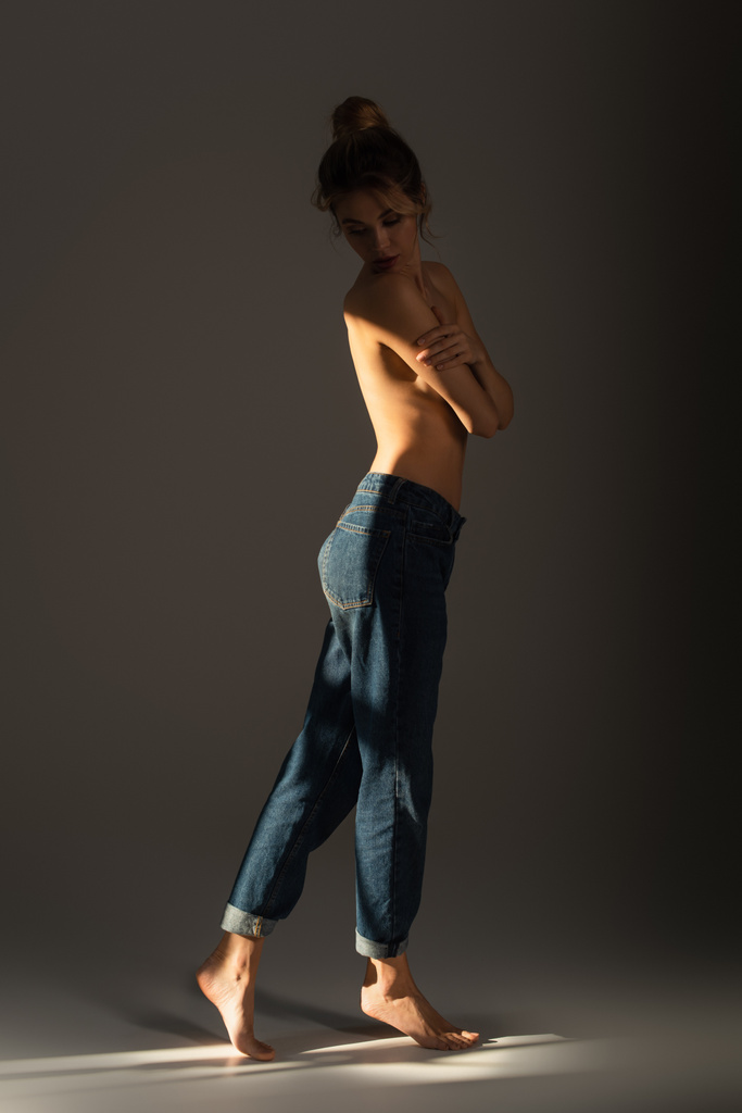 full length of half nude barefoot woman in jeans covering bust with crossed arms on grey background with lighting - Photo, Image