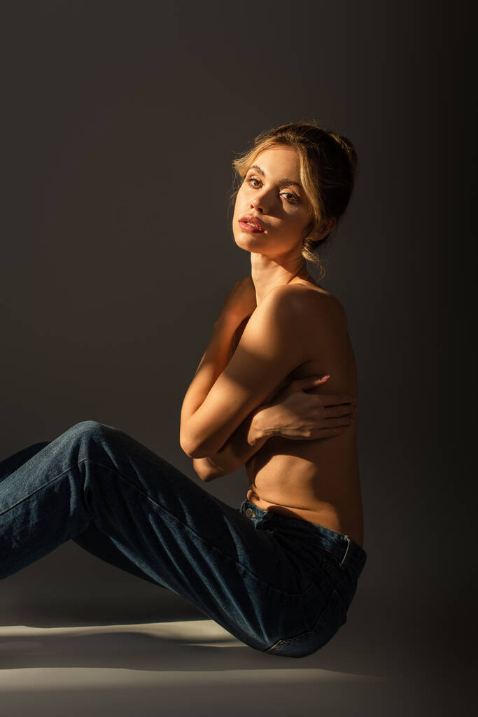 pretty half nude woman in jeans sitting with crossed arms and looking at camera on grey background with lighting - Photo, Image