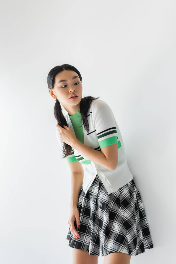 Hübsches asiatisches Modell in kariertem Rock und abgeschnittener Strickjacke auf grauem Hintergrund  - Foto, Bild