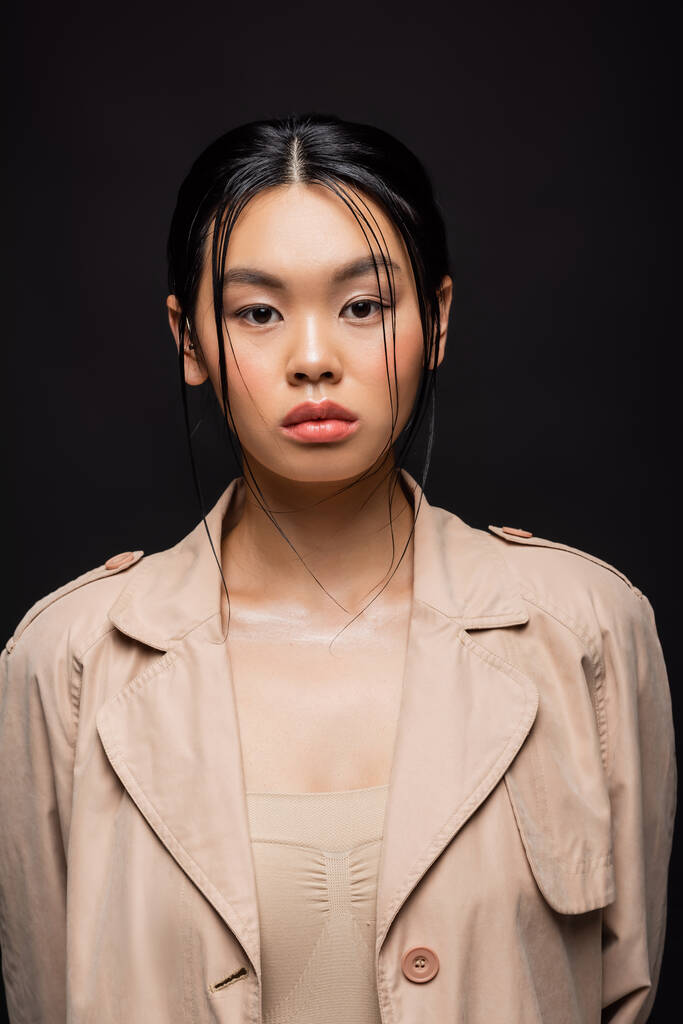 Portrait of asian woman in trench coat looking at camera isolated on black  - Photo, Image