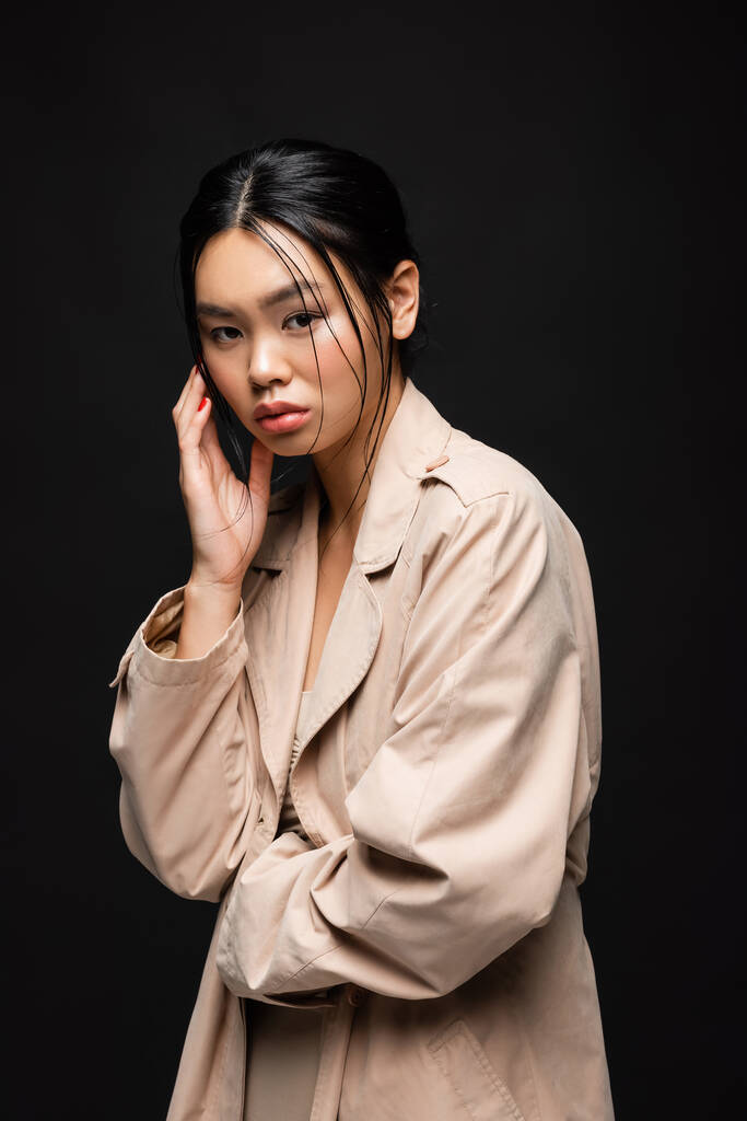 Fiatal ázsiai modell árok kabát nézi kamera elszigetelt fekete  - Fotó, kép