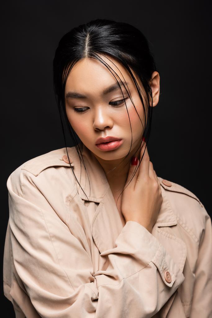 Portrét krásného asijského modelu v béžové zákopové srsti odvrací izolované na černé  - Fotografie, Obrázek