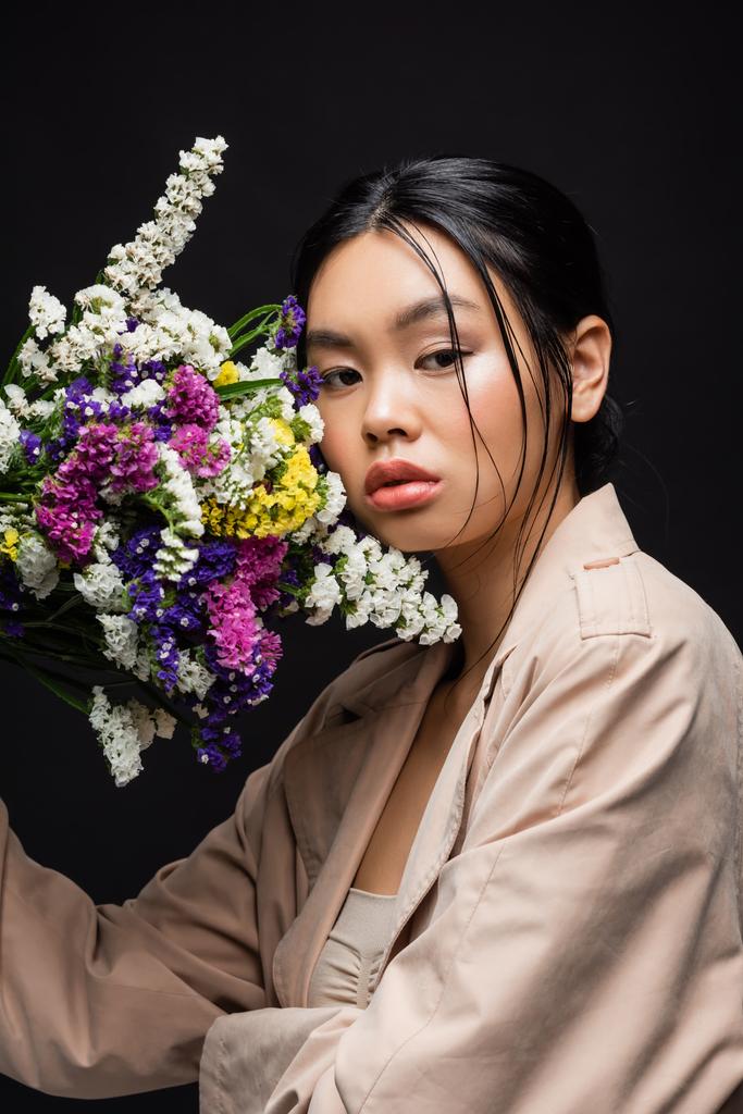 Молодая азиатская модель в плаще держа полевые цветы возле лица изолированы на черном  - Фото, изображение