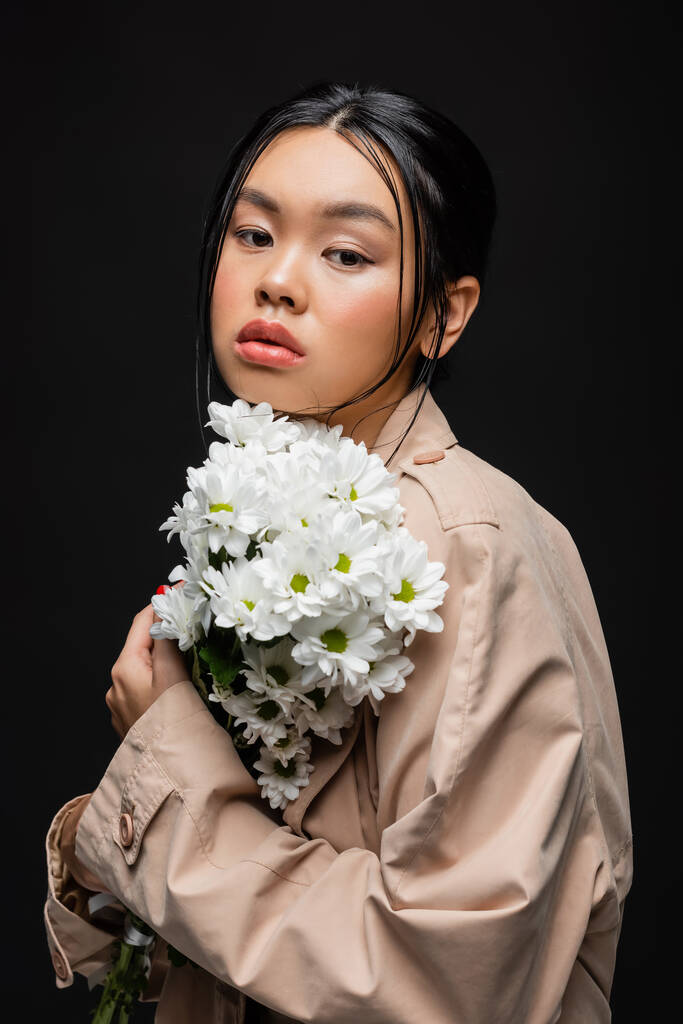黒に隔離された白い菊を保持トレンチコートのスタイリッシュなアジアの女性  - 写真・画像