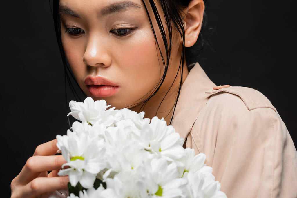 Portrait of asian woman in beige trench coat holding chrysanthemums isolated on black  - Photo, Image