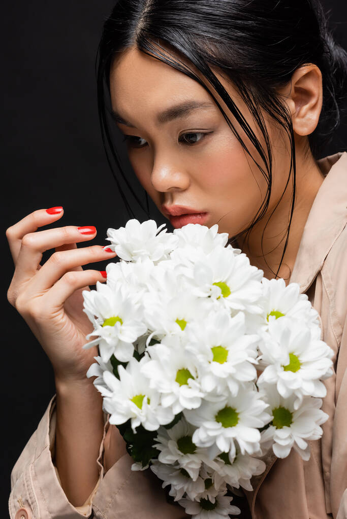 Stylish asian woman in trench coat posing with bouquet of chrysanthemums isolated on black  - Photo, Image