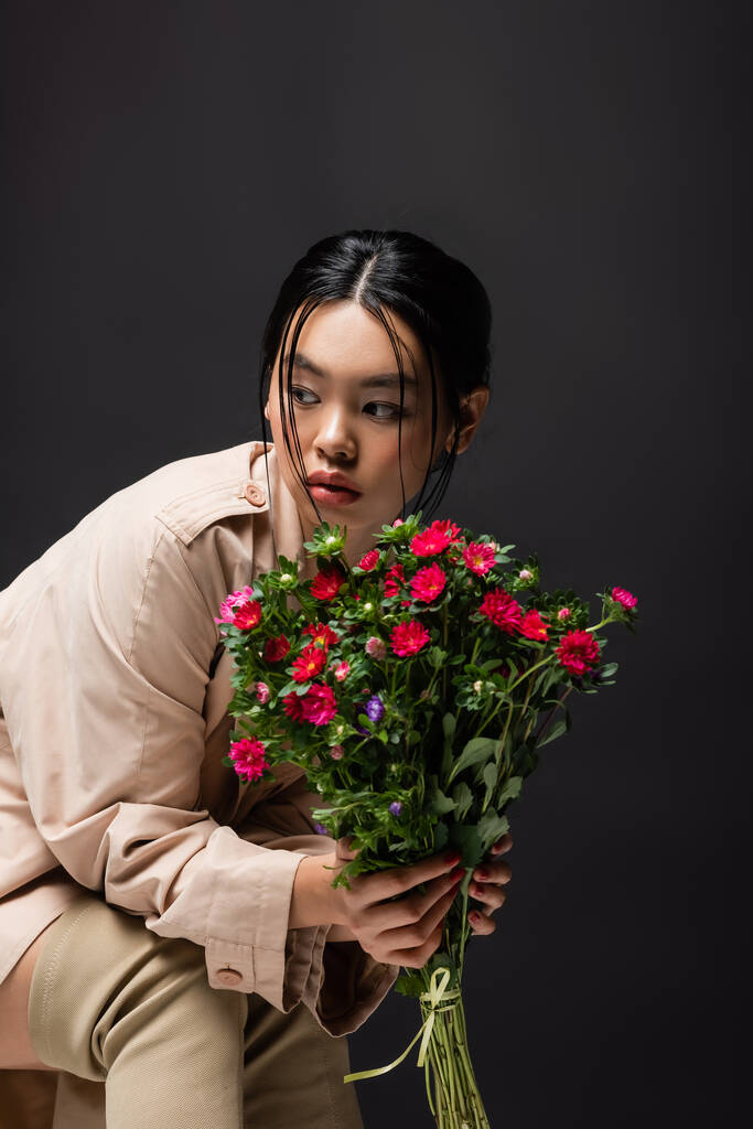 Trendy asian woman in boots and trench coat holding colorful flowers isolated on black  - Photo, Image