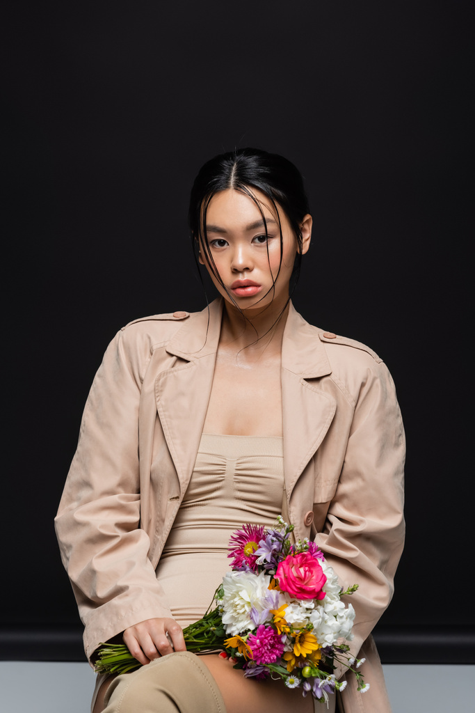 Modelo asiático na moda em casaco de trincheira segurando flores e olhando para a câmera no fundo preto  - Foto, Imagem