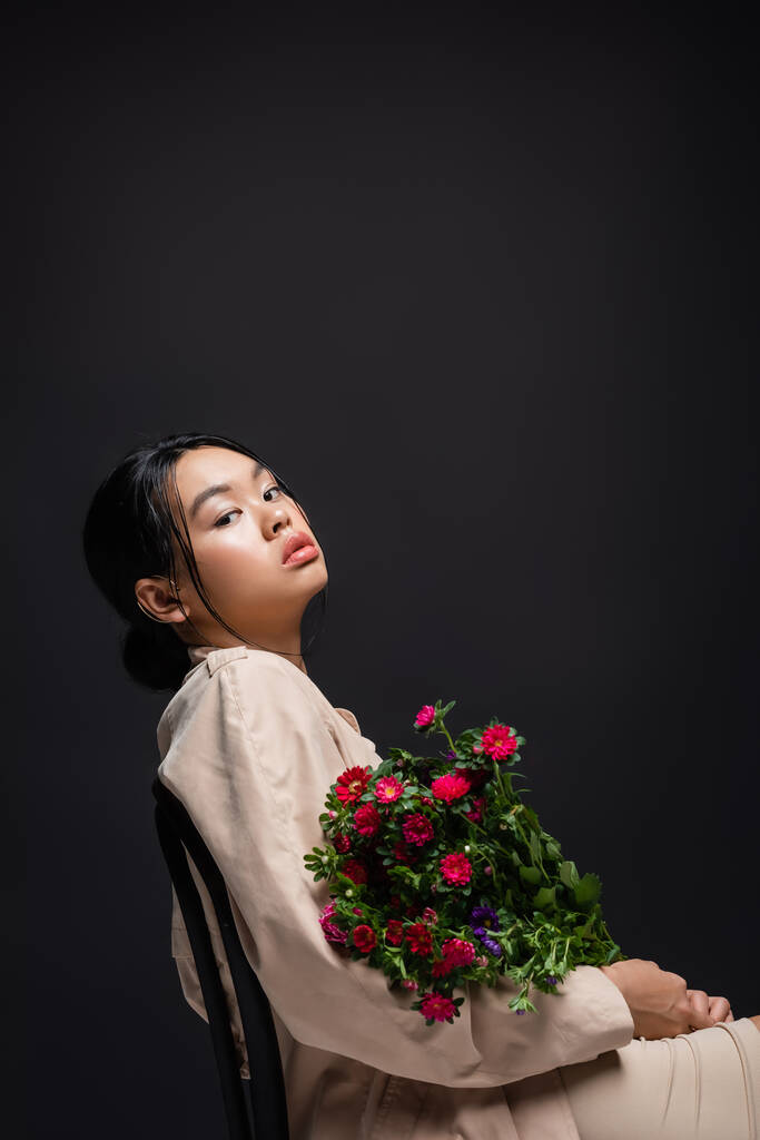 Stylish asian woman in trench coat and beige dress holding flowers while sitting on chair isolated on black  - Photo, Image