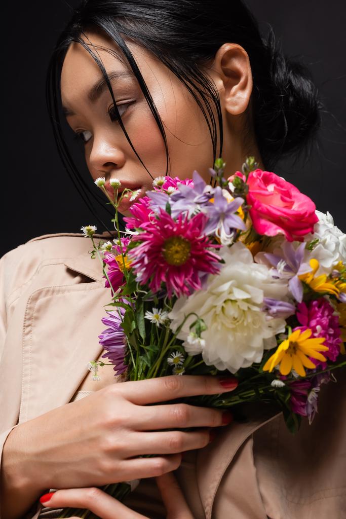 Portrait of asian model in trench coat holding colorful bouquet isolated on black  - Photo, Image