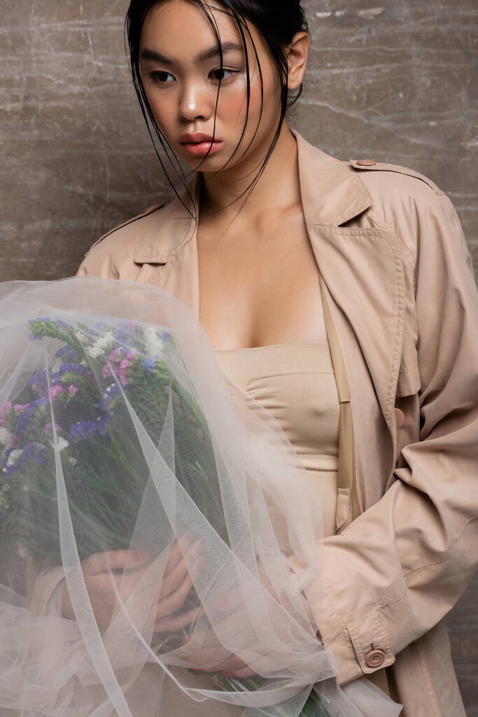 Сексуальная азиатка в плаще держит букет под вуалем на абстрактном коричневом фоне  - Фото, изображение
