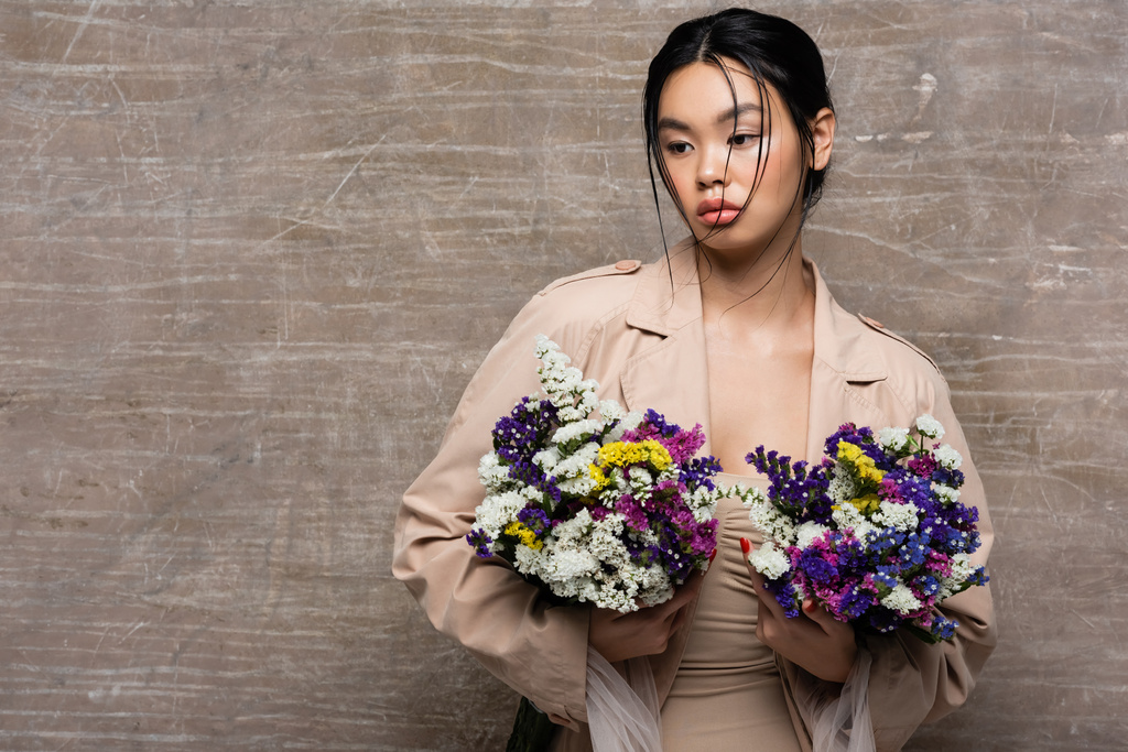 Jolie modèle asiatique en trench coat beige tenant des fleurs sauvages sur fond brun abstrait - Photo, image