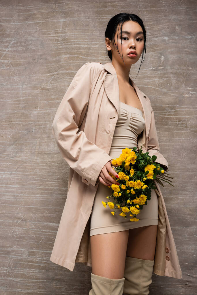 スタイリッシュなアジアの女性のドレスとブーツを保持黄色の花で抽象的な茶色の背景  - 写真・画像