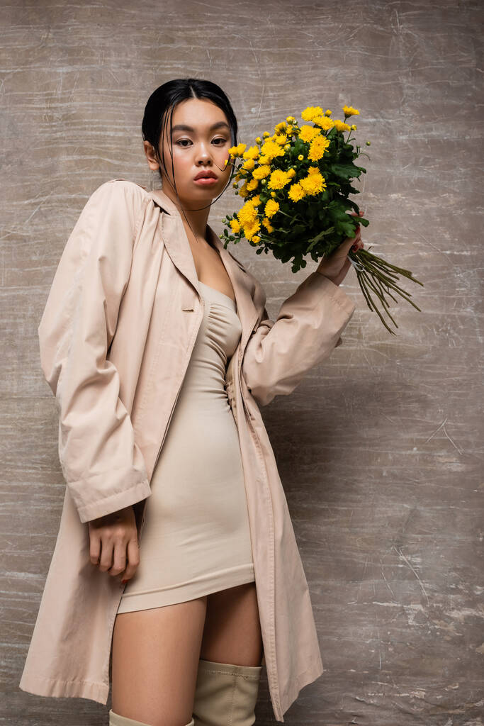 Low angle view of stylish asian woman in trench coat holding bouquet and looking at camera on abstract brown background  - Photo, Image