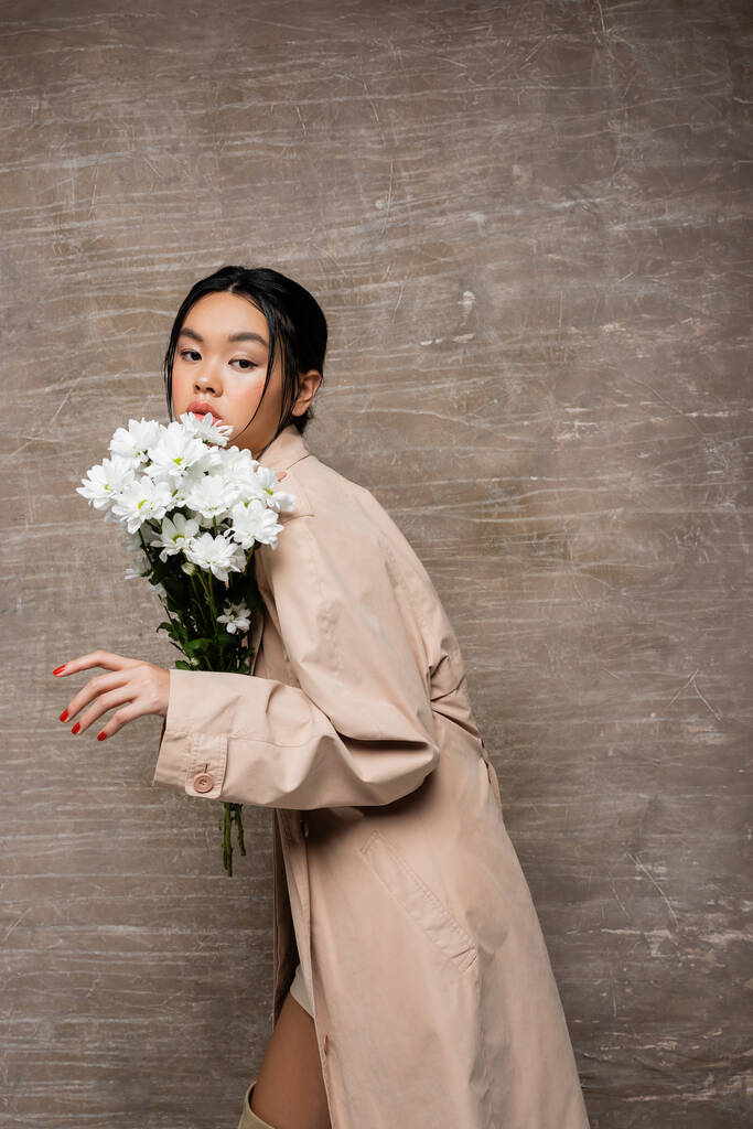 Pretty asian woman in trench coat posing with white chrysanthemums on abstract brown background  - Photo, Image