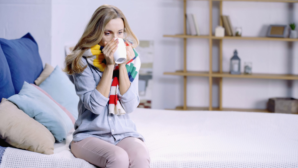 zieke vrouw in kleurrijke sjaal drinken thee en zitten op bed thuis - Foto, afbeelding