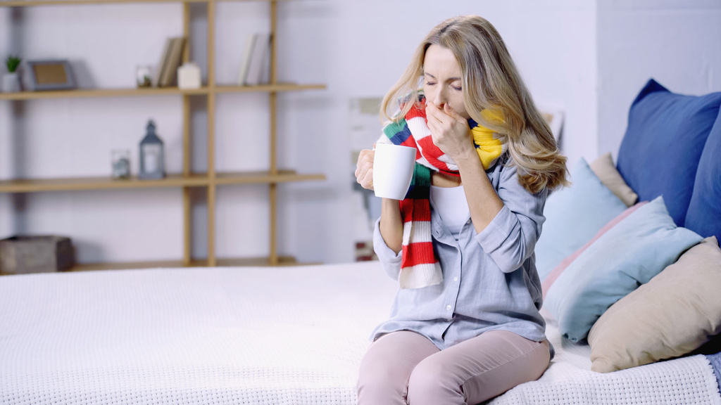 sick woman in colorful scarf drinking tea and coughing while sitting on bed at home - Photo, Image