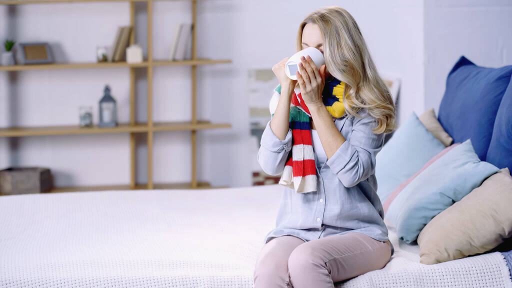 diseased woman in colorful scarf drinking tea and sitting on bed at home  - Photo, Image