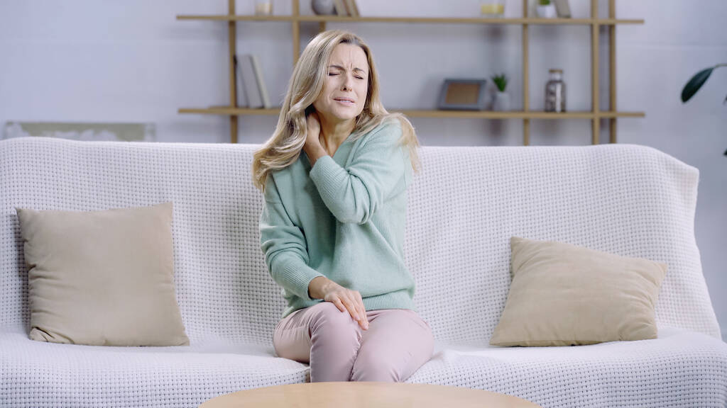 Frustrierte Frau mit Nackenschmerzen, während sie auf der Couch im modernen Wohnzimmer sitzt  - Foto, Bild
