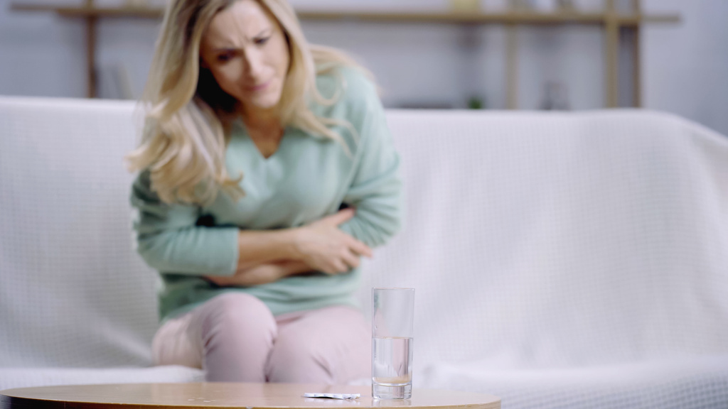 donna bionda che soffre di mal di stomaco vicino al bicchiere d'acqua e bottiglia con pillole sul tavolino  - Foto, immagini