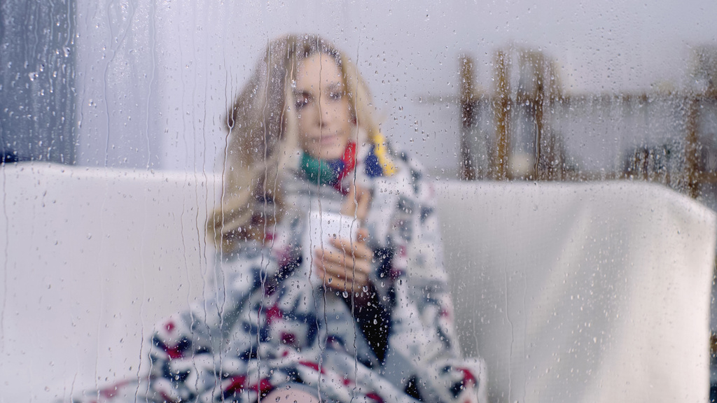 Kranke Frau sitzt unter bunter Decke und hält Tasse Tee hinter feuchtem Fenster mit Regentropfen  - Foto, Bild