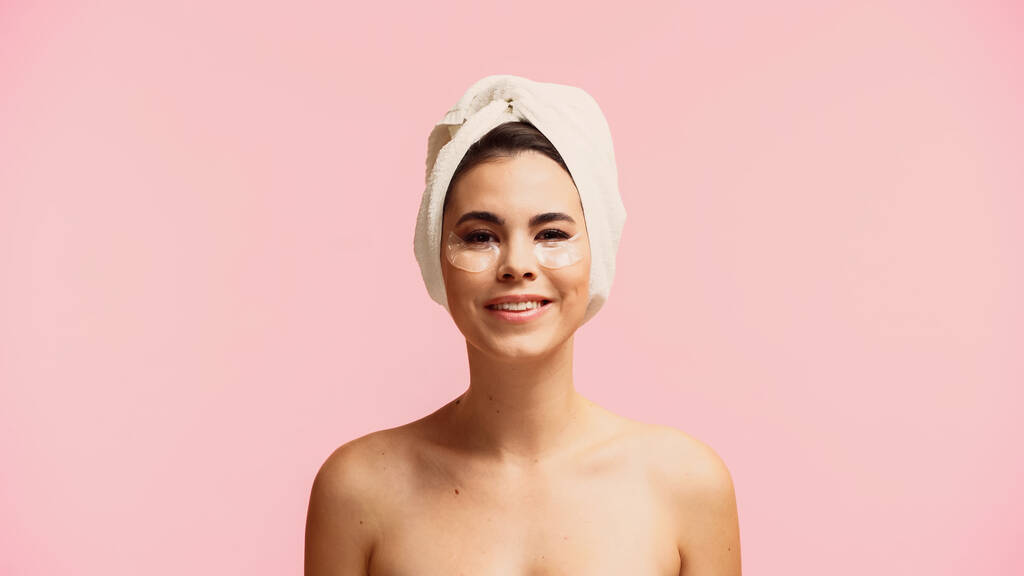mujer joven positiva con hombros desnudos y parches para los ojos hidratantes aislados en rosa  - Foto, imagen