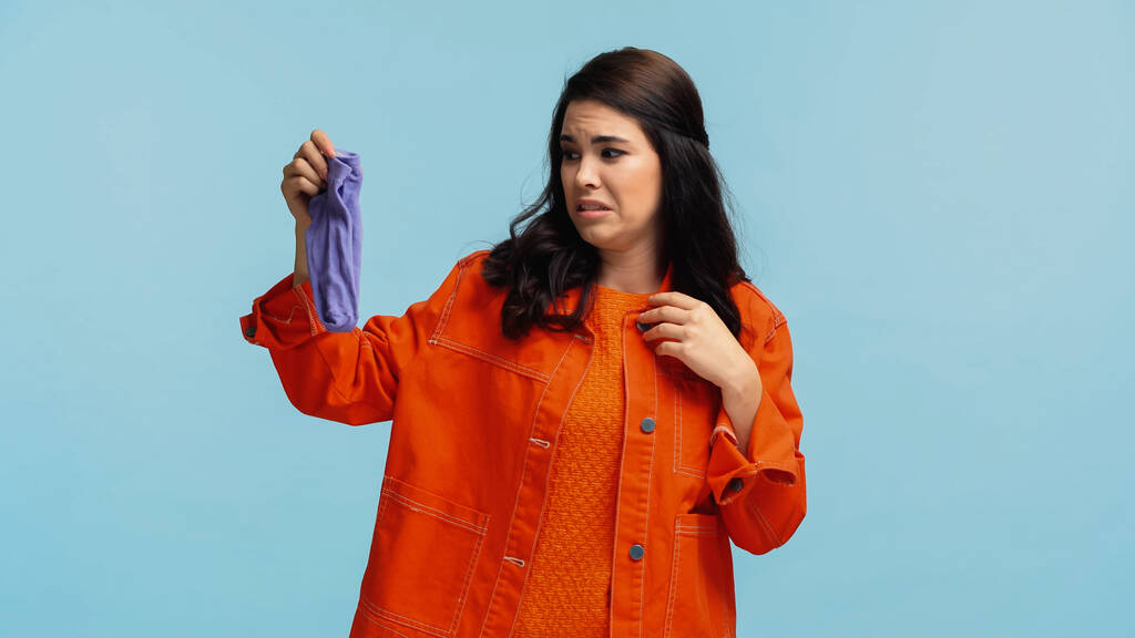 młoda i zdegustowana kobieta w pomarańczowej kurtce jeansowej trzymająca śmierdzące skarpetki odizolowane na niebiesko  - Zdjęcie, obraz