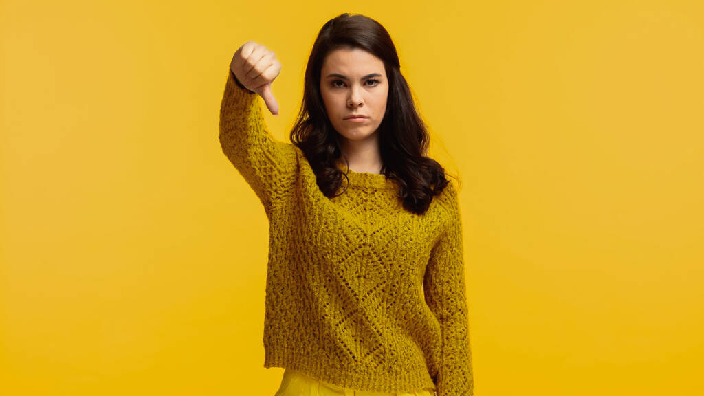 unpleased brunette woman in sweater showing thumb down isolated on yellow - Фото, изображение