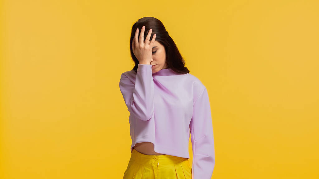 skeptische junge Frau in lila Sweatshirt stehend und Kopf auf gelbem Grund angefasst - Foto, Bild