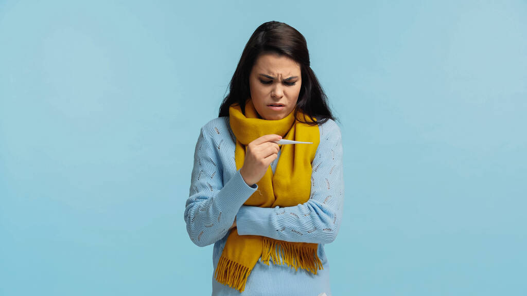 mujer triste y enferma en bufanda sosteniendo termómetro electrónico aislado en azul - Foto, imagen
