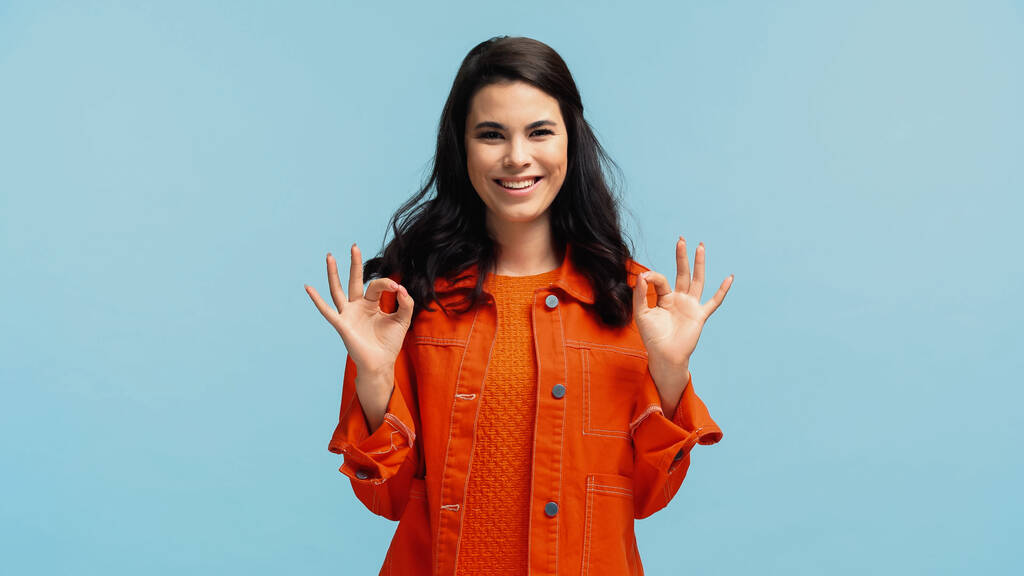 happy young woman in orange jacket showing ok isolated on blue - Photo, Image