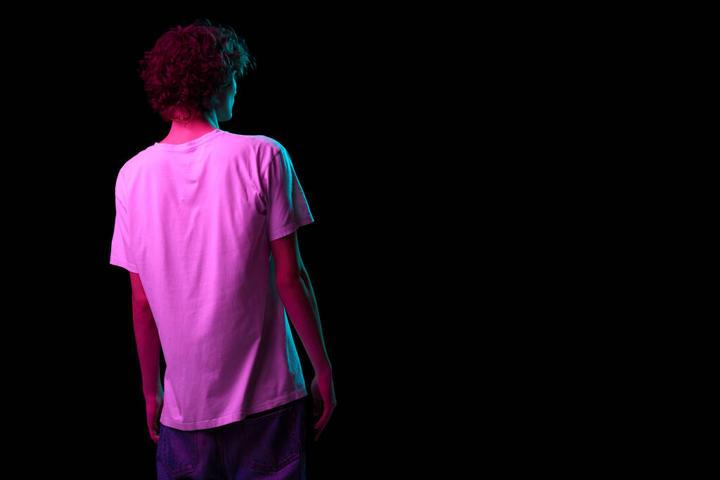 Mannelijk silhouet. Achteraanzicht van jonge harige man, student in witte tee geïsoleerd op donkere achtergrond in neon licht. Concept van emoties, gezichtsuitdrukking, jeugd, aspiratie, verkoop en reclame - Foto, afbeelding