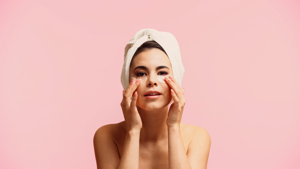 Nuori nainen paljain hartioin ja pyyhe päähän soveltamalla kosteuttava silmä laikkuja eristetty vaaleanpunainen  - Valokuva, kuva
