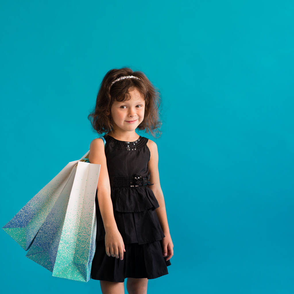 Dítě dívka v černých šatech s papírovými taškami na jednoduchém pozadí. Obchod pro slevy - Fotografie, Obrázek