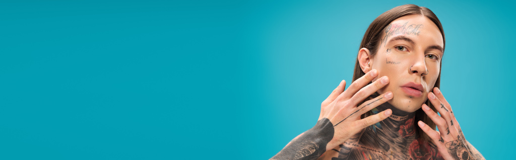 νεαρός άνδρας με τατουάζ αγγίζοντας λείο πρόσωπο απομονωμένο σε μπλε, banner - Φωτογραφία, εικόνα