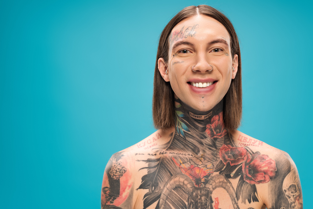 vrolijke jongeman met tattoos lachend en kijkend naar camera geïsoleerd op blauw - Foto, afbeelding