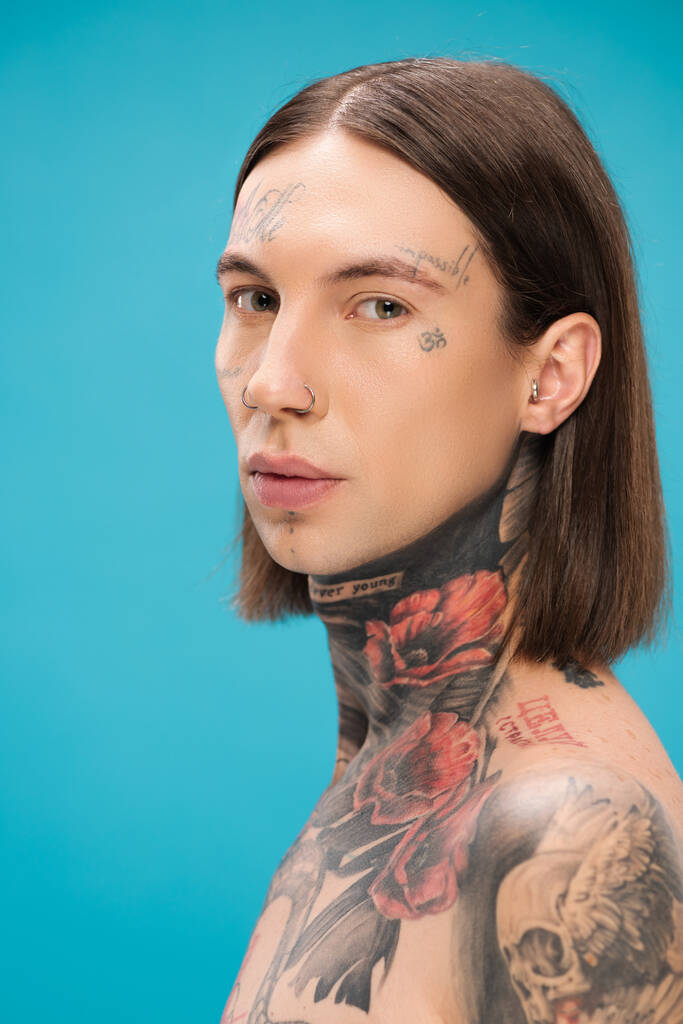 mladý a propíchnutý muž s tetováním při pohledu na kameru izolované na modré - Fotografie, Obrázek