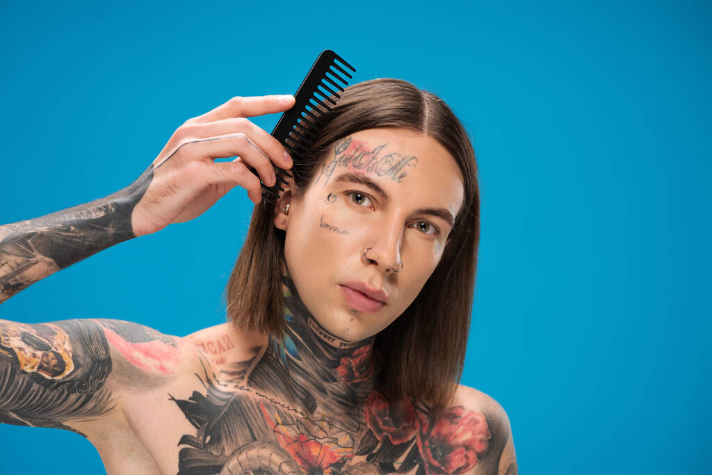 νέος και τρυπημένος άντρας με τατουάζ που βουρτσίζει τα μαλλιά του απομονωμένα στο μπλε - Φωτογραφία, εικόνα