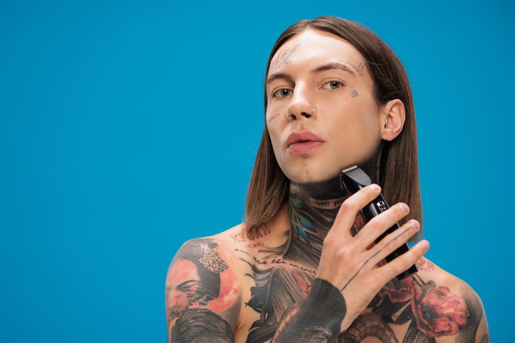 jovem e perfurado homem com tatuagens de barbear com lâmina elétrica isolada em azul - Foto, Imagem