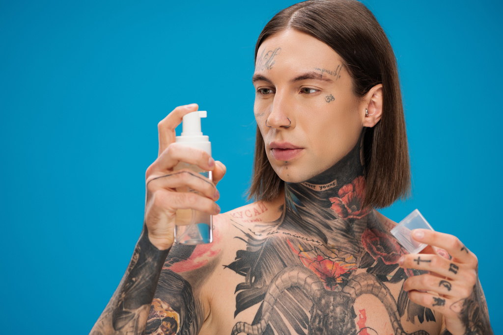 young and tattooed man looking at bottle with cleansing foam isolated on blue - Photo, Image