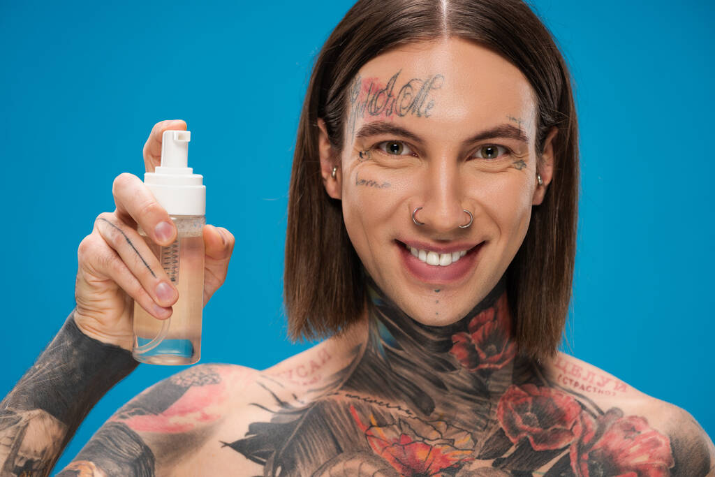 onnellinen ja tatuoitu mies tilalla pullo puhdistava vaahto eristetty sininen - Valokuva, kuva