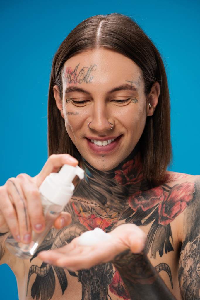 uomo sorridente e tatuato in bottiglia con schiuma detergente isolata su blu - Foto, immagini