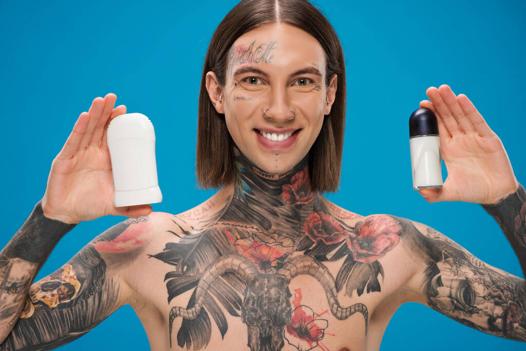 χαρούμενος και με τατουάζ άντρας που κρατάει διαφορετικά αποσμητικά απομονωμένα στο μπλε - Φωτογραφία, εικόνα
